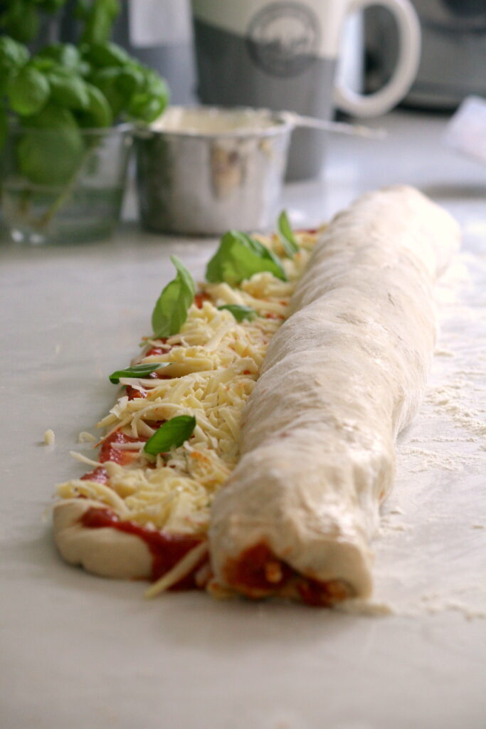 white pizza dough rolled into a long roll on a white surface with basil growing behind