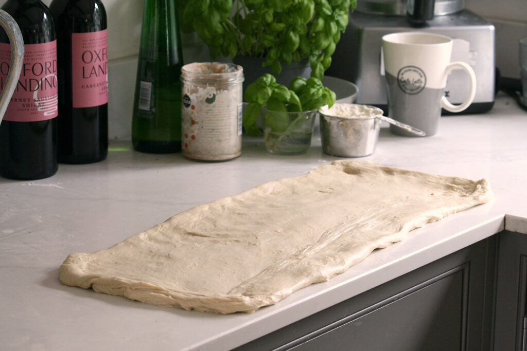white pizza dough rolled out on a white surface with basil growing behind
