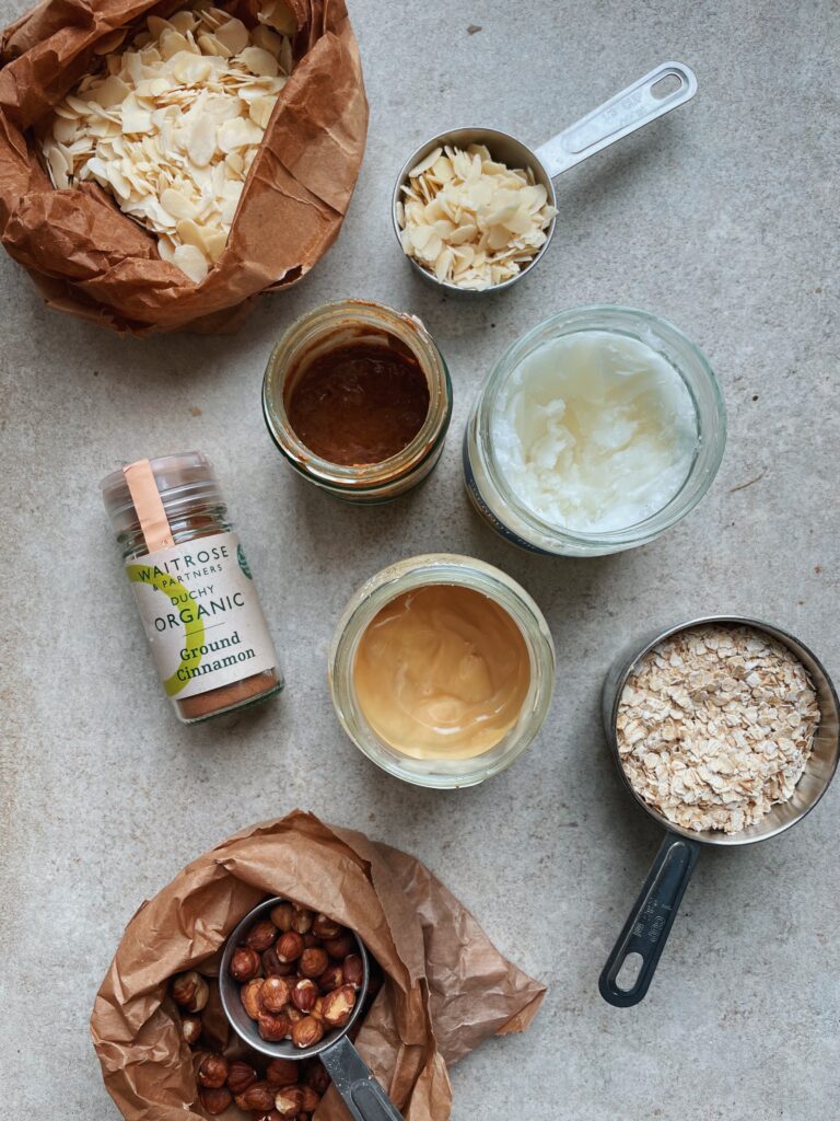 granola ingredients laid out on a grey tile background in a top down shot. Metal cups and glass jars with oats, nuts and nut butters